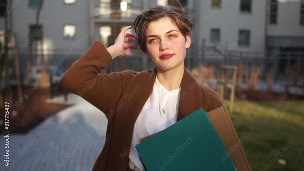 Short-haired girl in a brown coat holds a folder with papers in her hands. Young businesswoman in the courtyard of an office building at sunset Short-haired girl in a brown coat holds a folder with papers in her hands. Young businesswoman in the courtyard of an office building at sunset
