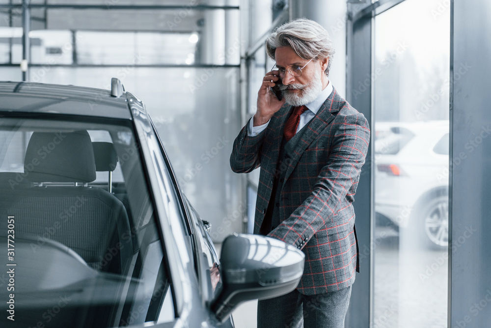 Senior businessman in suit and tie with gray hair and beard standing indoors with phone near car