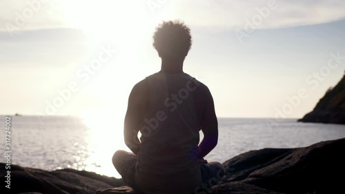Back view of man sitting in lotus position and meditating at sunset by the sea