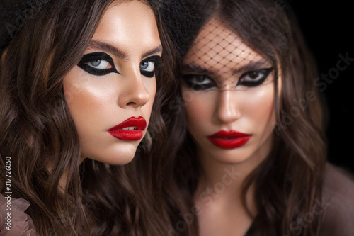 Art photo of two attractive girls with graphic cat eye makeup and red lips. One model is closer than another. Twins.