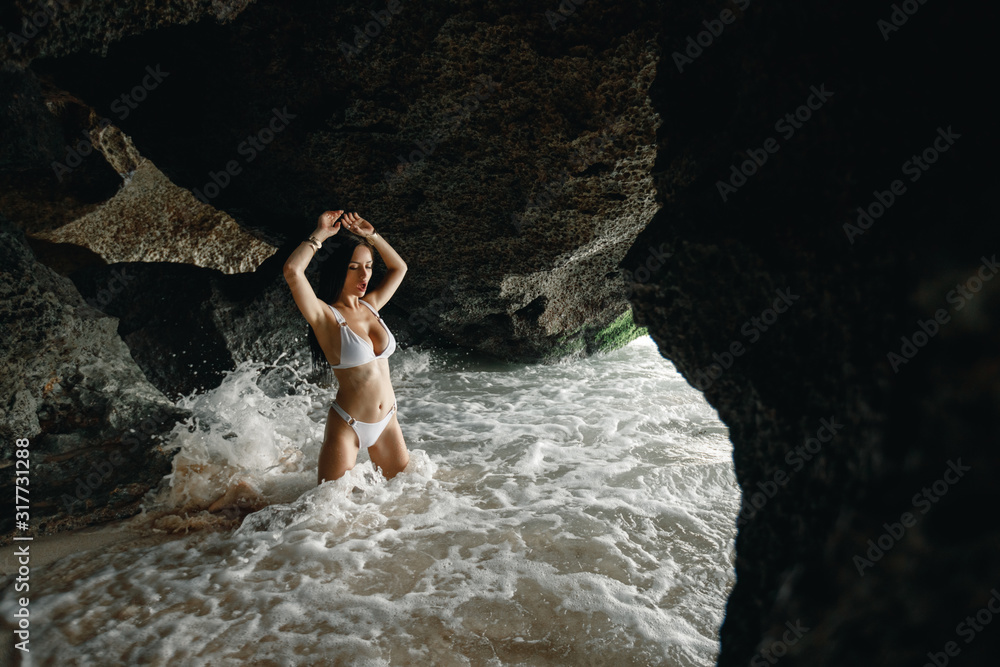 Brunette girl in clear water on a hidden beach in Bali, Indonesia luxury lifestyle. Amazing young woman is resting on the beach. Tanned girl in a white bathing suit. Portrait of a model near a rock