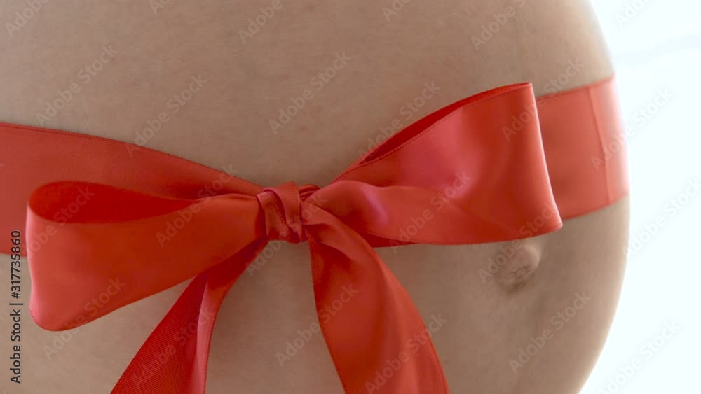 Closeup red ribbon with bow decorating pretty tummy of pregnant woman, maternity Closeup red ribbon with bow decorating pretty tummy of pregnant woman, maternity