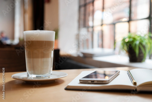 Glass of latte on the wooden table with smartphone and notebook for writing and planning. Loft indoors with big window for coffee time. Break at wotk.