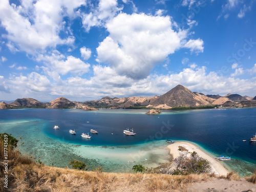 A view from the top of a cliff of Kelor Island on the bay and white sand beach, Komodo National Park, Indonesia. There is another island in the back. There are boats anchored to the shore. Happiness