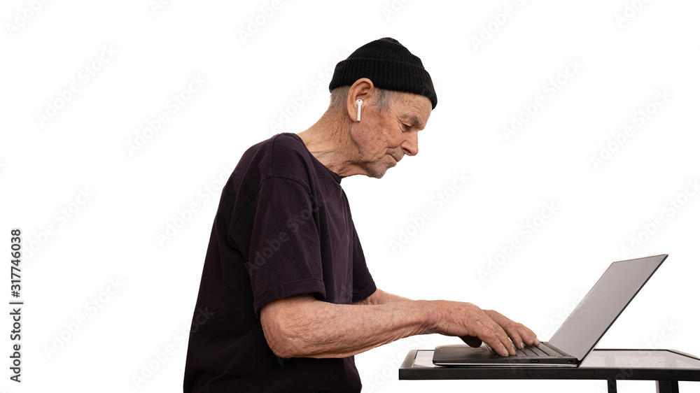 Serious old man in black hat, t-shirt and white wireless headphones, senior white hacker typing on the keyboard, works at a laptop, isolated over white background Serious old man in black hat, t-shirt and white wireless headphones, senior white hacker typing on the keyboard, works at a laptop, isolated over white background