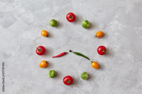 Moving colored vegetables make up a clock with moving arrows from chili pepper on a gray concrete background. Stop motion animation.