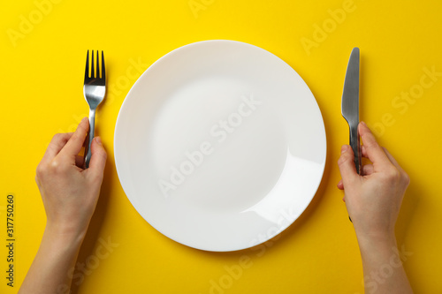 Obraz na plátně Female hands hold fork and knife on yellow background with plate, top view