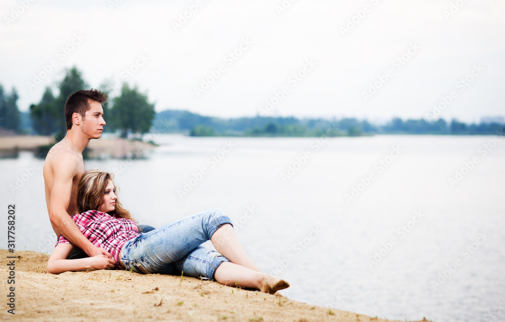 Young beautiful loving couple having rest on river shore on summer day with green nature at background. Love, relationships, dating, flirt and attraction concept