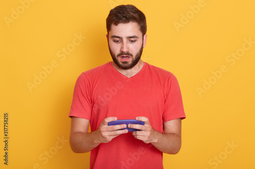 Horizontal shot of young bearded man dressses casual clothes posing isolated over yellow background, handsome male sing mobile phone, typing sms message, having astonished facial expression.