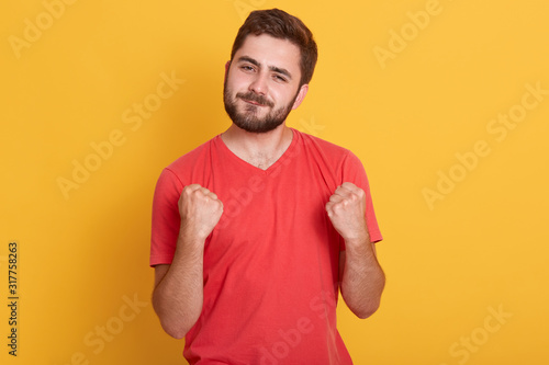 Portrait of grinning excited bearded man clenching fists near chest and looking directly at camera, guy celebrating success or victory, model standing isoalted over yellow background. People concept.