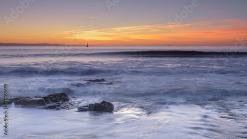 Waves rolling over rocks on the shoreline, while the sun sets over the horizon