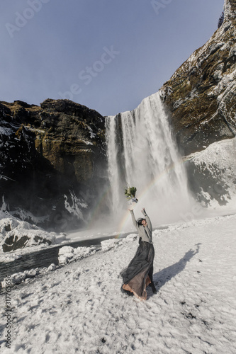 young beautiful wedding bride in black clothes stands near a waterfall in Iceland