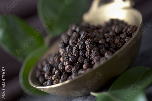 Brass spoon with black peppercorns and leaves on textile, close up photo