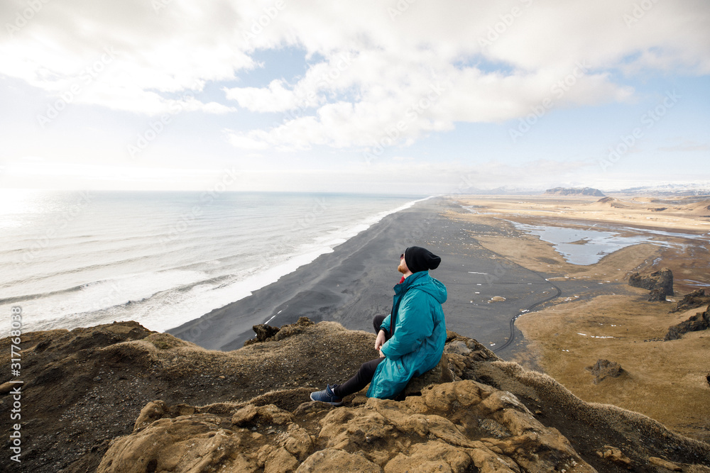 male traveler in green clothes and sitting near a cliff view of a black beach and the Atlantic Ocean in Iceland male traveler in green clothes and sitting near a cliff view of a black beach and the Atlantic Ocean in Iceland
