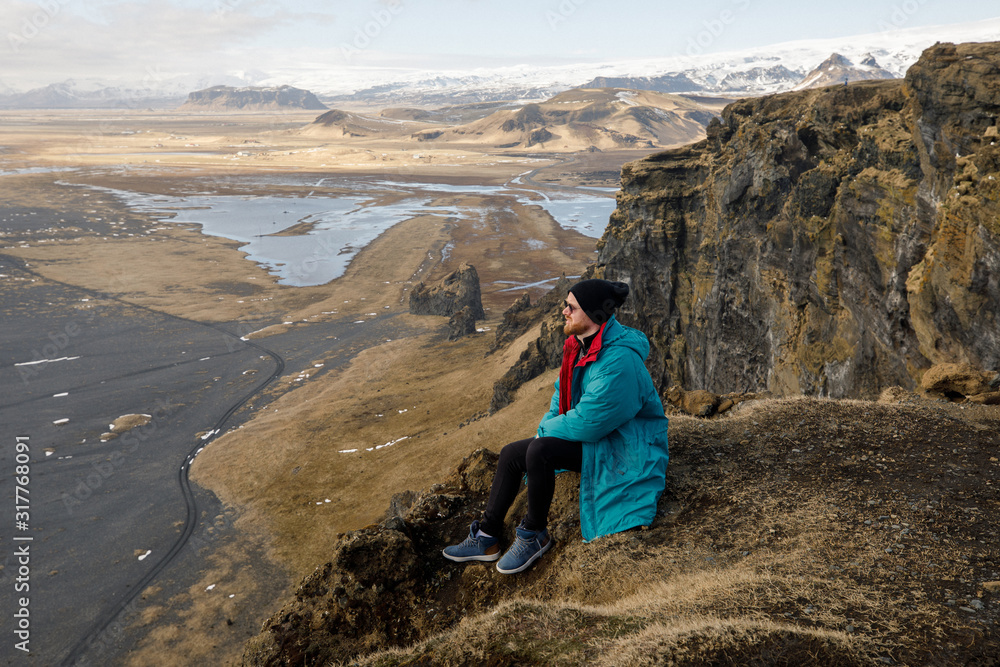 male traveler in green clothes and  sitting near a cliff view of a black beach and the Atlantic Ocean in Iceland