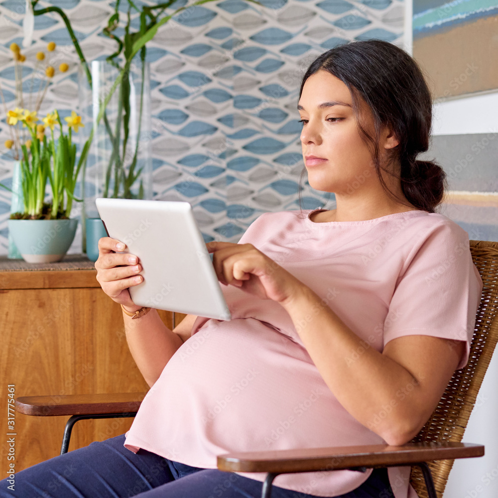 Young pregnant woman sitting on a chair using her tablet Young pregnant woman sitting on a chair using her tablet