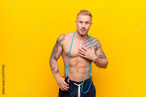 young strong blonde man feeling shocked, astonished and surprised, with hand on chest and open mouth, saying who, me? with a jump rope