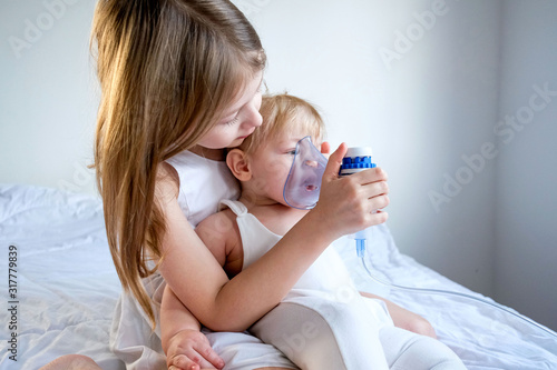 The children got sick. The older sister takes care of her brother and holds a gmu mask for inhalation. Medicine at home. stylish white room photo