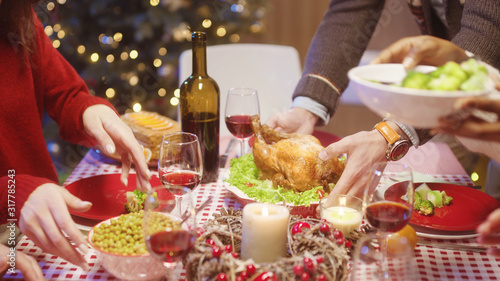 Christmas dinner. Hands of man placing hot delicious turkey on dining table. Friends passing by food on table celebrating holiday together.