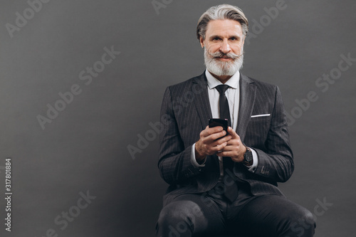 Business and fashionable bearded mature man in a gray suit sitting and using smartphone on the grey background.