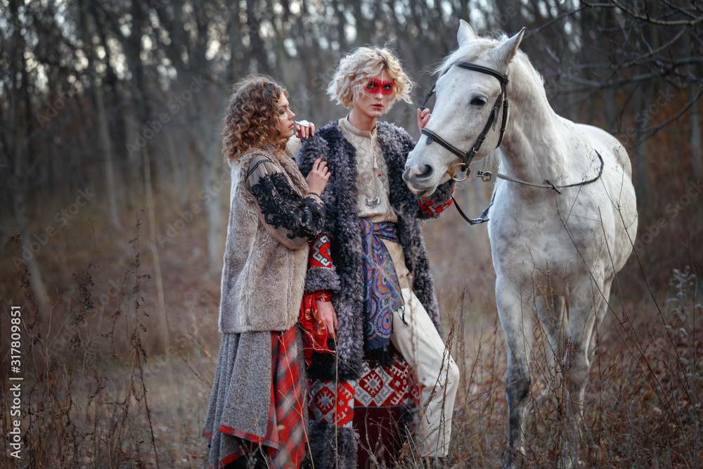A young attractive man with curly blond hair with an ethnic ritual pattern on the face and ethnic clothes of the East European Slavs with a bride girl holds a horse under a knot in a light suit. A young attractive man with curly blond hair with an ethnic ritual pattern on the face and ethnic clothes of the East European Slavs with a bride girl holds a horse under a knot in a light suit.