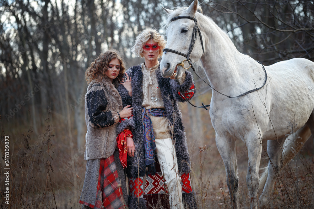 A young attractive man with curly blond hair with an ethnic ritual pattern on the face and ethnic clothes of the East European Slavs with a bride girl holds a horse under a knot in a light suit.
