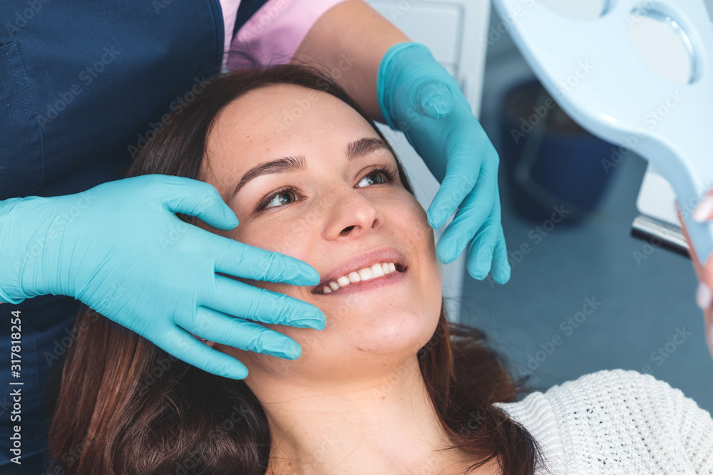 Portrait of happy female patient sitting in dentist chair, smiling and examining her teeth in the mirror, dental surgeon showing the work with her hands in blue gloves, health care concept