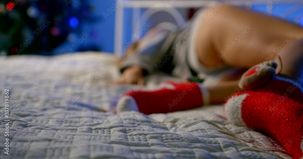 Close-up of a young girl in her underpants lying in bed in a Christmas decorated room, with a Christmas tree and glowing garlands behind her, and red Christmas socks on her feet. Close-up of a young girl in her underpants lying in bed in a Christmas decorated room, with a Christmas tree and glowing garlands behind her, and red Christmas socks on her feet.