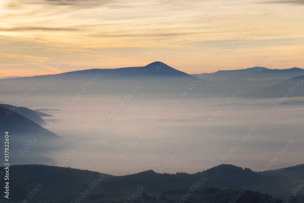 Valley filled with soft mist, distant silhouettes of impressive mountains and amazing colors of vivid morning sky