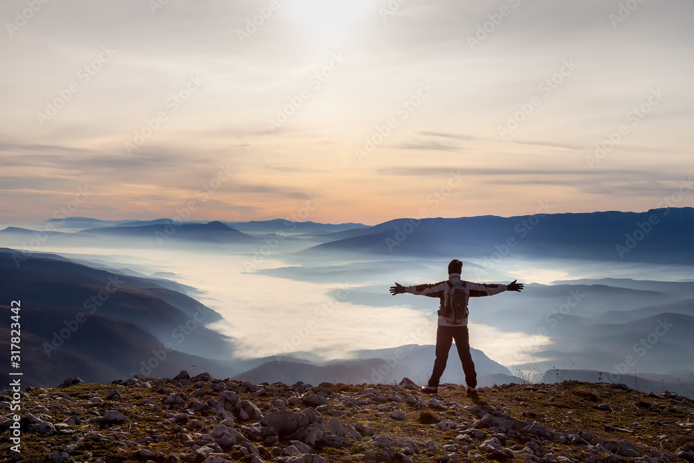 Mountain hiker standing at the edge of mountain peak with hands in the air and looking at a valley filled with thick fog during colorful sunrise
