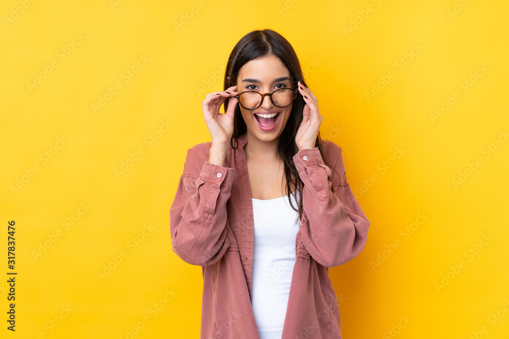 Young brunette woman over isolated yellow background with glasses and surprised Young brunette woman over isolated yellow background with glasses and surprised