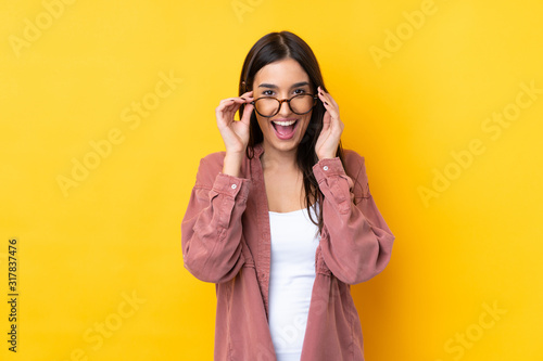 Young brunette woman over isolated yellow background with glasses and surprised