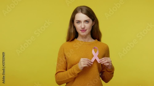 Smiling cute girl with pleasant kind friendly face expression holding pink ribbon, sign of breast cancer prevention, disease awareness and healthcare. indoor studio shot isolated on yellow background