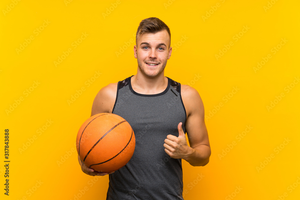 Young handsome blonde man holding a basket ball over isolated yellow background with thumbs up because something good has happened