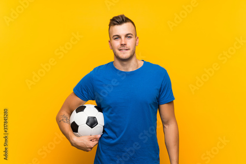 Young handsome blonde man holding a soccer ball over isolated yellow background
