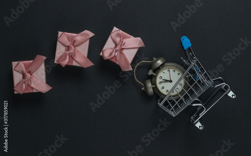 Retro alarm clock, shopping trolley, gift boxes with bow on black background. 11:55 am. New Year, Christmas concept. Holiday Shopping. Top view