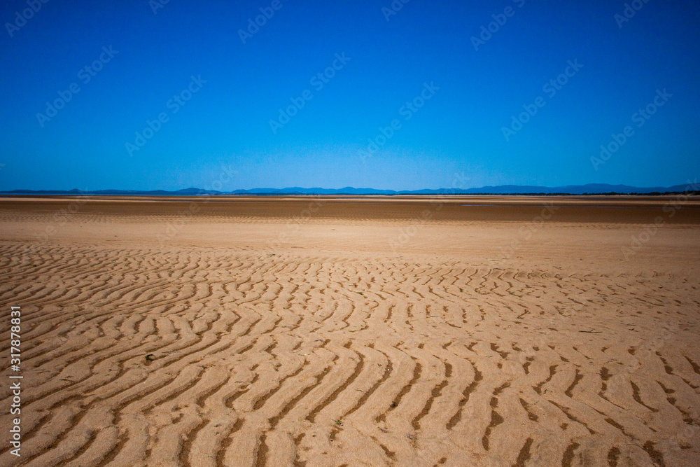 Fototapeta premium The coastal strip (coastline, beach) at low tide. A wide strip of desert sand to the horizon, a desert area, a wavy relief runs along it. Makey, Queensland, Australia.