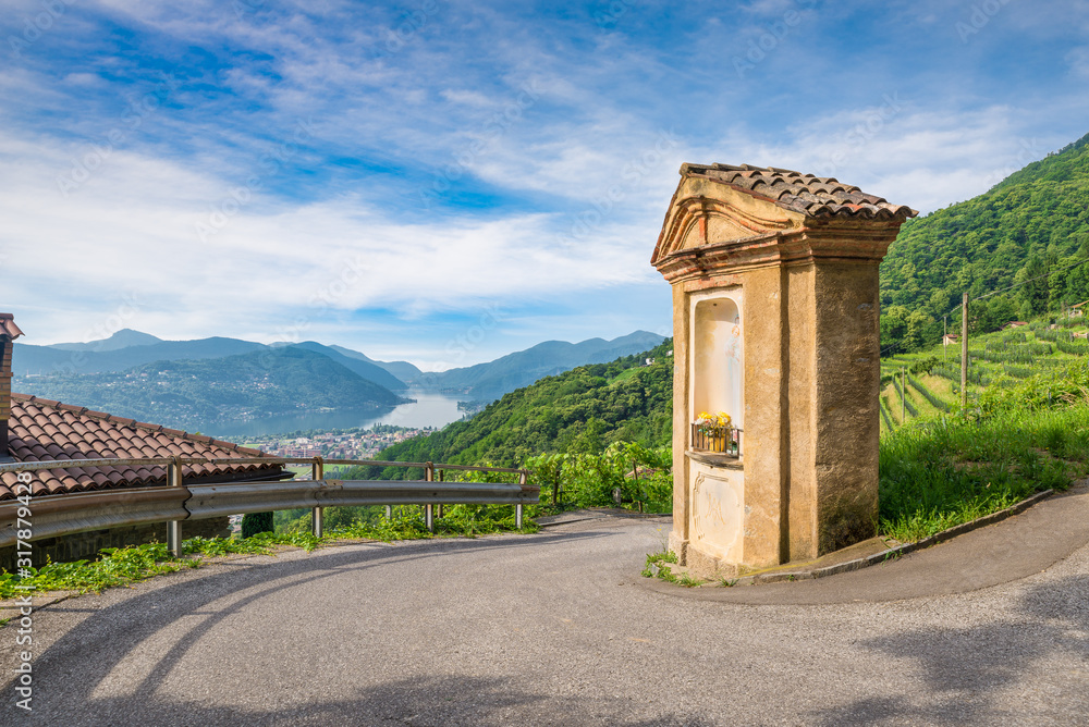 Scenic view of lake and mountains against blue sky. High angle view of lake Lugano and the town of Agno from the hills of Switzerland with a small votive chapel in the foreground