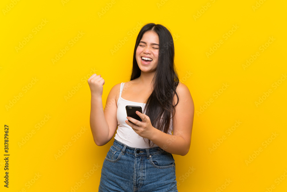 Young teenager Asian girl over isolated yellow background with phone in victory position Young teenager Asian girl over isolated yellow background with phone in victory position