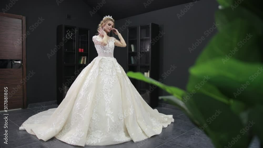 Young blonde bride in white wedding dress with crown poses for camera in room with black walls. Green leaves of houseplant are blurred in foreground Young blonde bride in white wedding dress with crown poses for camera in room with black walls. Green leaves of houseplant are blurred in foreground