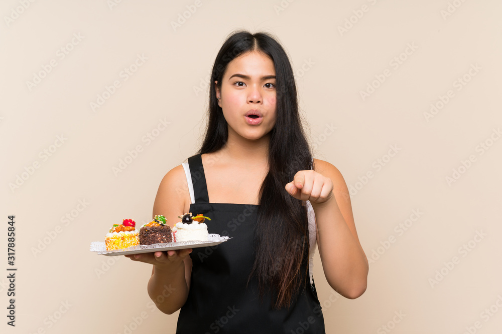 Young teenager Asian girl holding lots of different mini cakes over isolated background surprised and pointing front Young teenager Asian girl holding lots of different mini cakes over isolated background surprised and pointing front