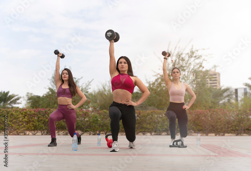 Three beautiful athletic brunette  women exercising in a park wearing tight active sports wear in a park with dumbbells and kettlebells 