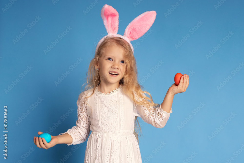 Portrait of a cheerful laughing girl in a white dress and pink rabbit ears holding Easter eggs in her hands. isolated on a blue background Portrait of a cheerful laughing girl in a white dress and pink rabbit ears holding Easter eggs in her hands. isolated on a blue background