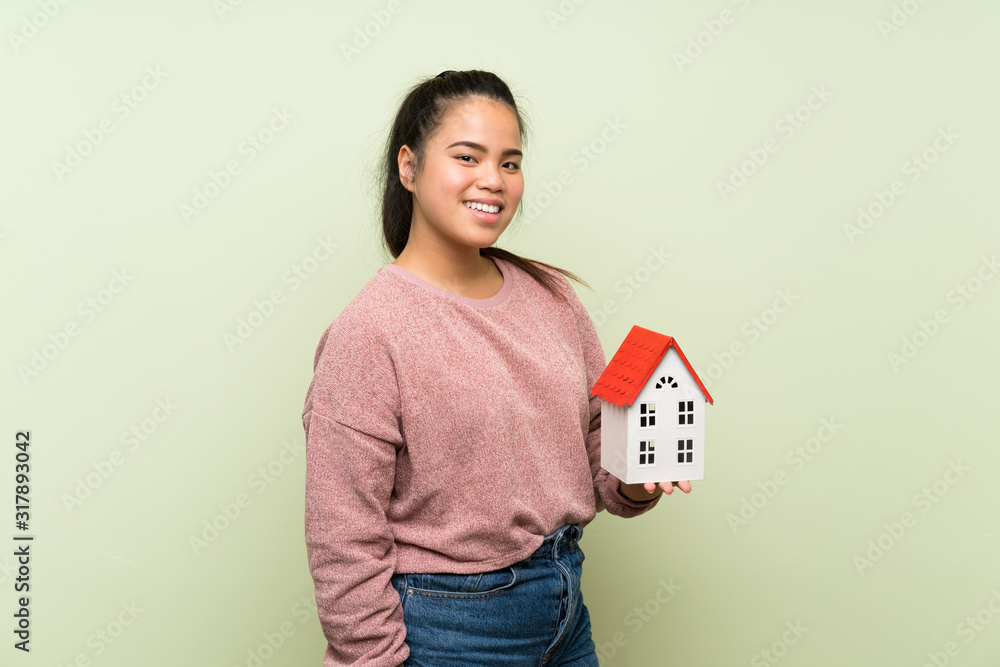 Young teenager Asian girl over isolated green background holding a little house Young teenager Asian girl over isolated green background holding a little house