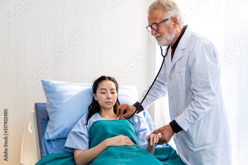 Senior professional Caucasian man examining Asian woman patient with stethoscope in the hospital room.