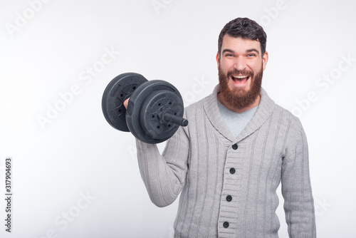 A young bearded man is smiling to the camera and lifting up a black dumbbell on white background.