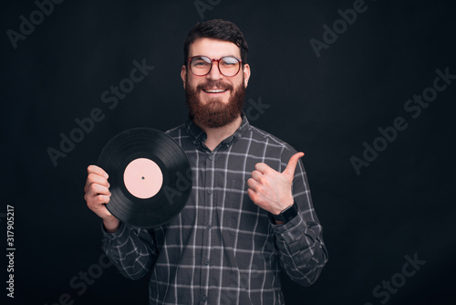 A bearded man holds a round vinyl record and shows a thumb up near black wall.