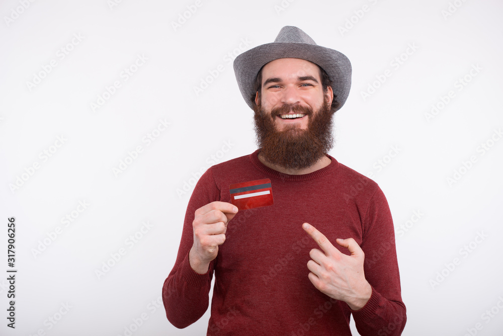 Bearded man is smiling and holding a credit card is pointing at it near a white wall. Bearded man is smiling and holding a credit card is pointing at it near a white wall.