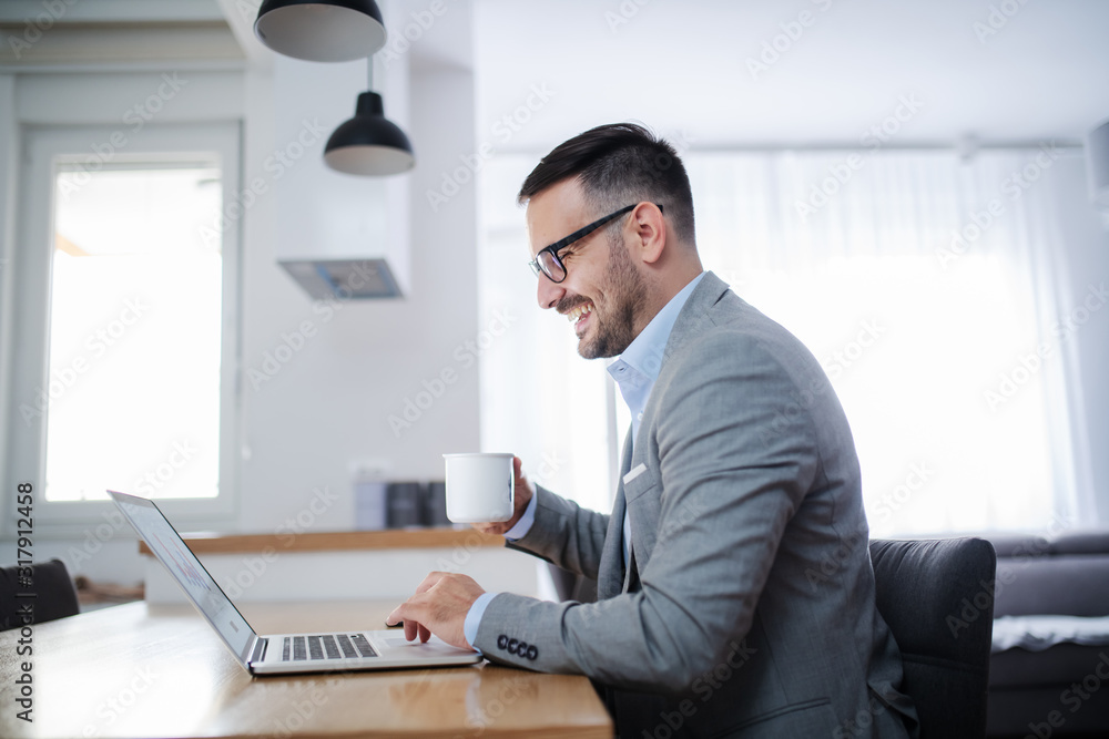 Side view of handsome unshaven caucasian businessman in suit and with eyeglasses sitting at dining table, using laptop and drinking morning coffee. Home interior.