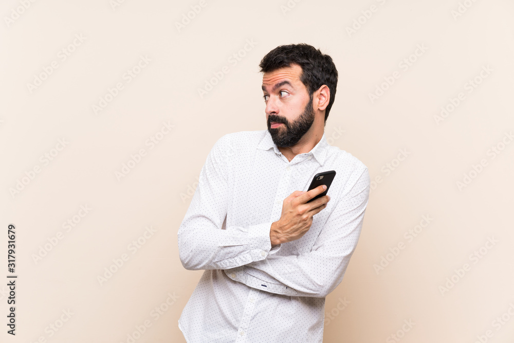 Young man with beard holding a mobile making doubts gesture while lifting the shoulders Young man with beard holding a mobile making doubts gesture while lifting the shoulders
